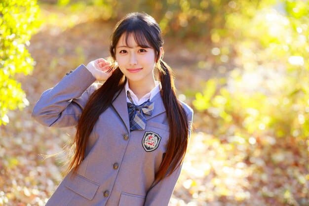 制服姿のツインテールの女性が落ち葉の屋外で立ち、左手を髪に添えて正面を見る