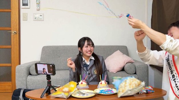 制服姿の女性がソファ前のテーブルで笑い、ケーキや菓子とスマホ撮影用の三脚が置かれる