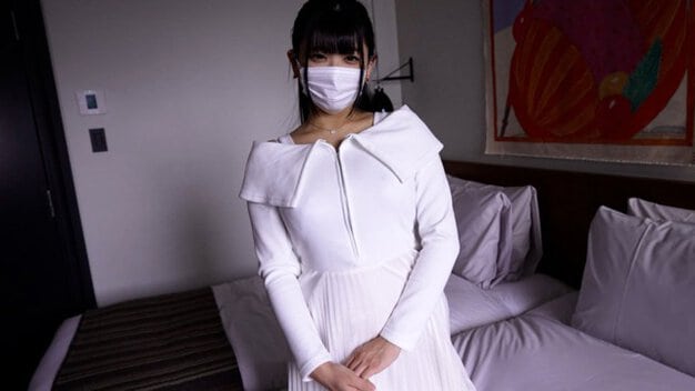 白い服とマスク姿の女性がベッドの前に立ち、両手を重ねてこちらを見る