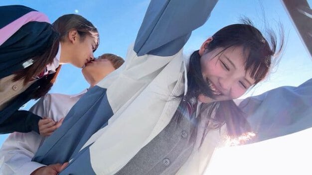 制服を着た黒髪の女性が男性とキスをし、もう一人の黒髪の女性が隣で笑いながら手を上げている。背景は青空。