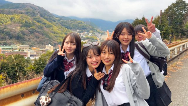 4人の女性と男性1人が道路に立ち、山を背景にピースサインをしている。4人は制服姿で、男性はメガネをかけている。左側の女性は黒いカバンを持ち、サングラスを頭にかけている。