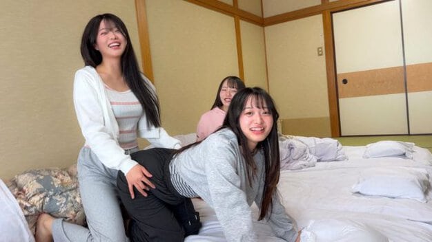 3人の女性がベッドの上にいる。1人が前かがみになり、もう1人が後ろからその尻に手を置いている。もう1人は後ろで座っている。全員が私服を着用している。