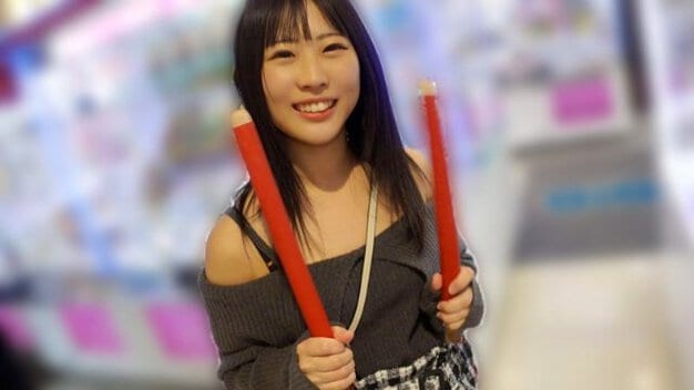 黒髪ロングヘアの女性がゲームセンター内で立っている。グレーのオフショルダーニットを着用し、両手で赤い太鼓のバチを握っている。背景にはぼやけたアーケードゲーム機が見える。女性は正面を向いて笑顔を見せている。