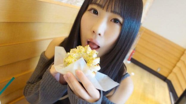 黒髪ロングヘアの女性が屋内で座っている。グレーのオフショルダーニットを着用し、両手で黄色いトッピングが乗ったクリーム系の菓子を持っている。口を開けて菓子を食べようとしている。背景は木目調の壁と床。